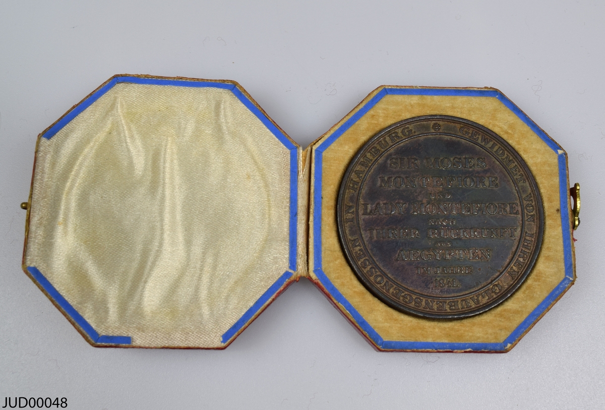 Medalj liggandes i pappask, som invändigt är klädd med siden och sammet. Medaljen är rikligt dekorerad med ett släktträd, vapensköld och hebreisk text på ena sidan samt flera namn på andra sidan.