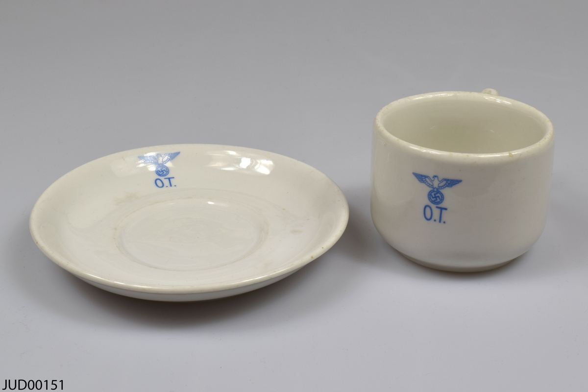 Kopp med tillhörande fat, tillverkade av vitt porslin. Både koppen och fatet är dekorerad med en nazistisk örn hållandes swastika och initialerna O.T i blått. Tillverkade i Norge.