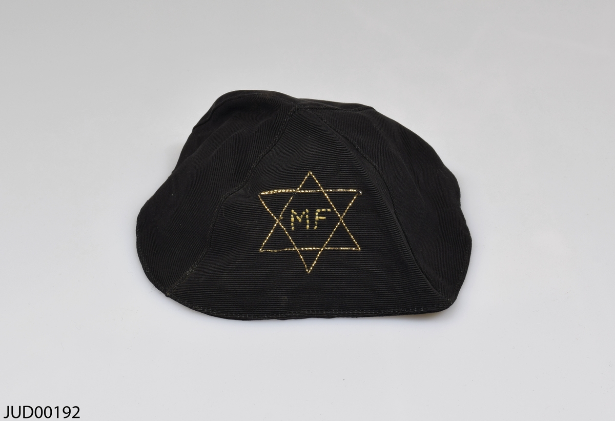 Kipa tillverkad av svart textil. Dekorerad med en davidsstjärna med initialerna MF inuti, broderade med guldfärgad tråd.