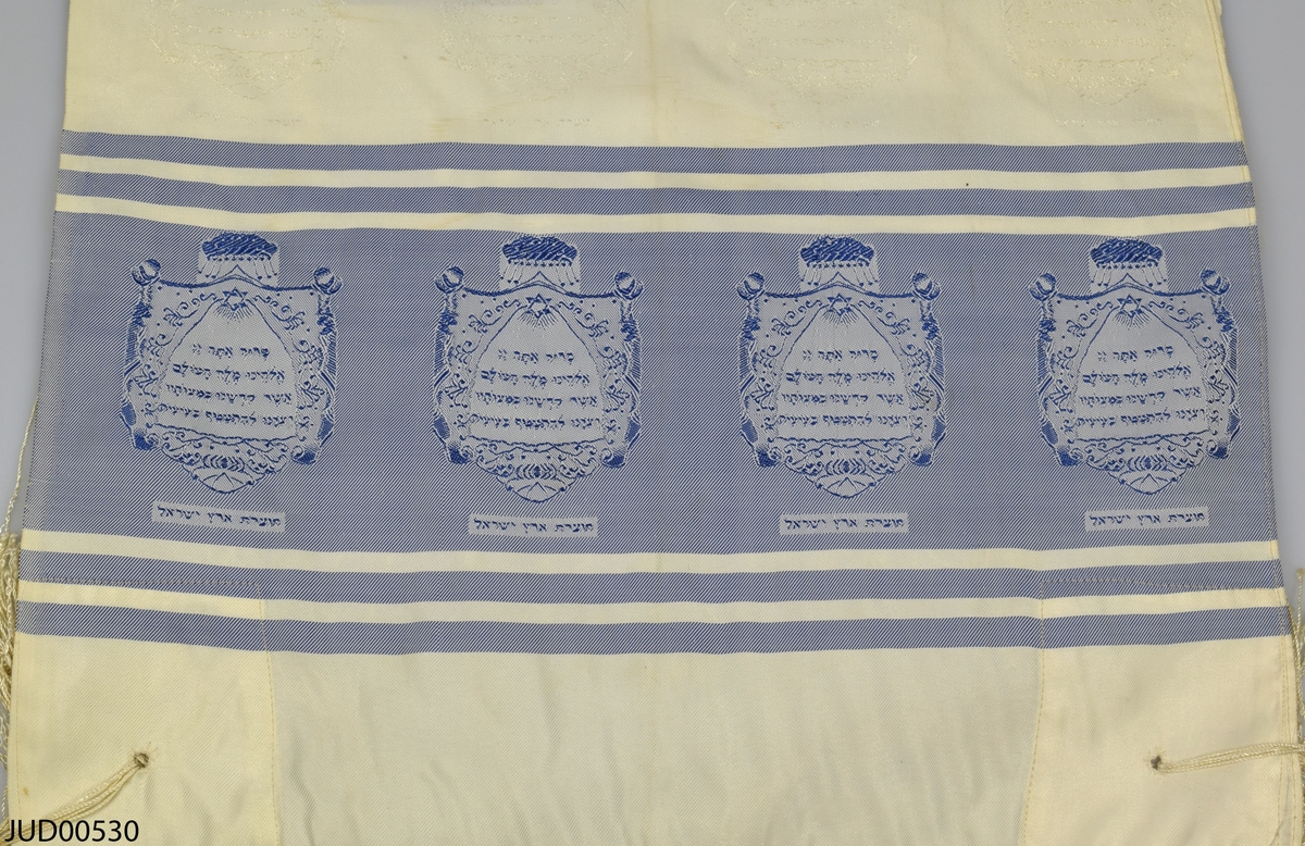 Bönesjal, troligtvis tillverkad av maskintillverkad satin. Invävd dekor i form av blå ränder med infällda sköldar i vilka hebreisk text är skriven. Likdana sköldar i vitt på det vita. I ena kanten finns ett band med hebreisk text.