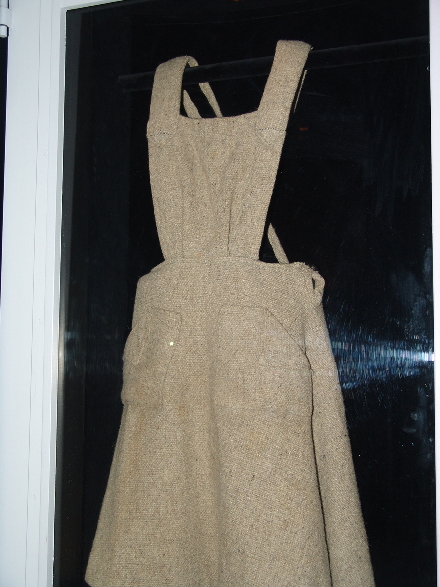 Grå klänning tillverkad av ett grovt tyg, troligtvis ull. Två fickor framtill och hängslen med knäppning i ryggen med knapp. Troligtvis sydd av en filt från koncentrationslägret Bergen-Belsen.