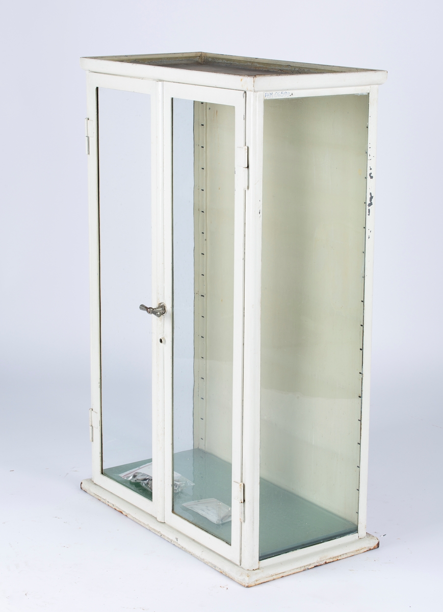 Rektangulært instrumentskap med fire glasshyller. Det er glass i høyre dør og i sidevegger. I den venstre døren mangler glasset.