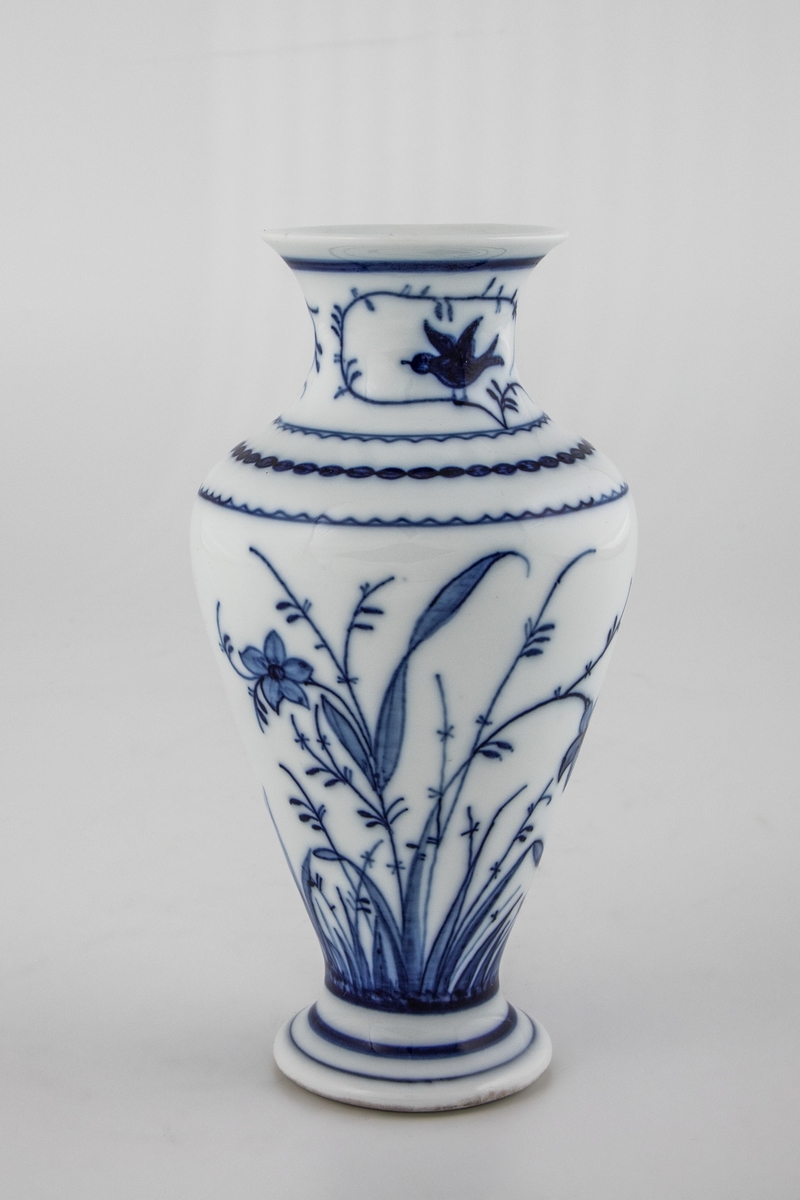 Vase i glasert porselen, dekorert med blomsterdekor i koboltblå underglasurmaling. Balusterformet koprus med utoverbrettet munningsrand, som hviler på en hevet fot.