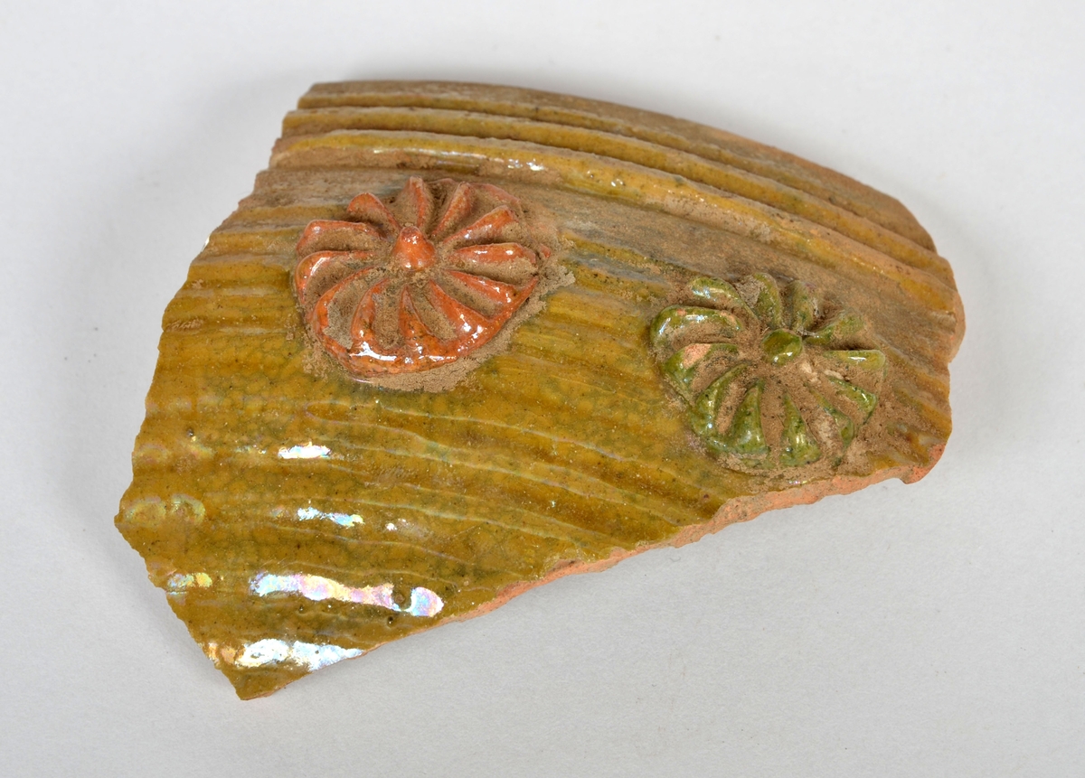 Keramikskärva. Del av kanten till en skål med gulgrön glasyr på både in- och utsida. Utsidan smyckad med två blomrosetter, en med röd och en med grön glasyr.