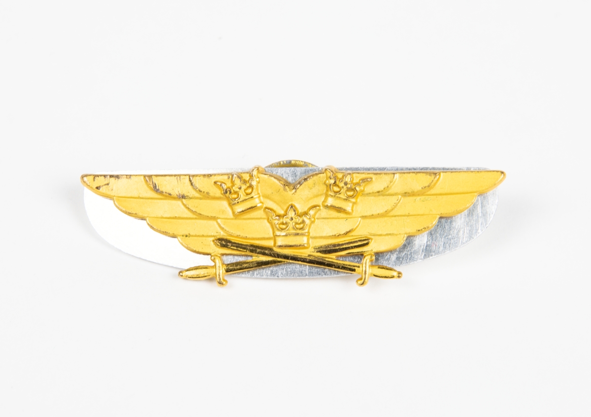 Flygförarmärke m/36 i gulmetall. Formad som ett vingpar med tre kronor över korsade svärd. Baksidan präglad med nummer: "6838".
Medföljer en skiva i vitmetall, för placering mellan flygförarmärket och dess skruvfäste.