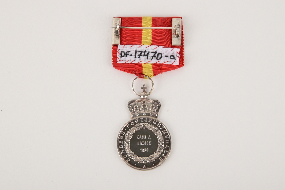 Et eksemplar av "Kongens fortjenstmedalje" i originalt etui. 

Sirkulær medalje med ordensbånd, oppbevart i rektangulært etui. Etuiet har motiv av en krone i sølv på lokket.