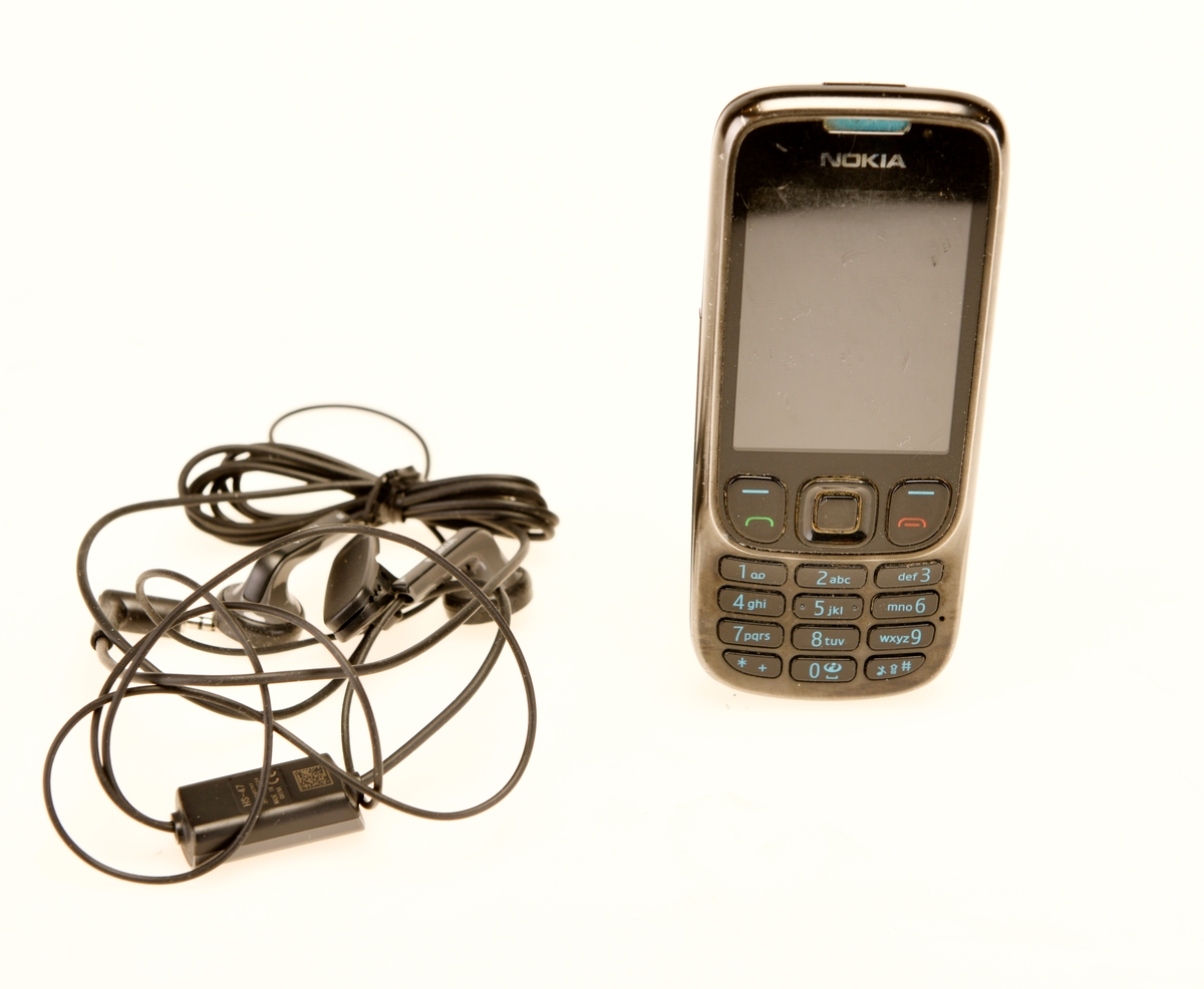 Svart mobilteleton av modellen NOKIA 6303 med tilhørende hodetelefoner HS-47 i svart.