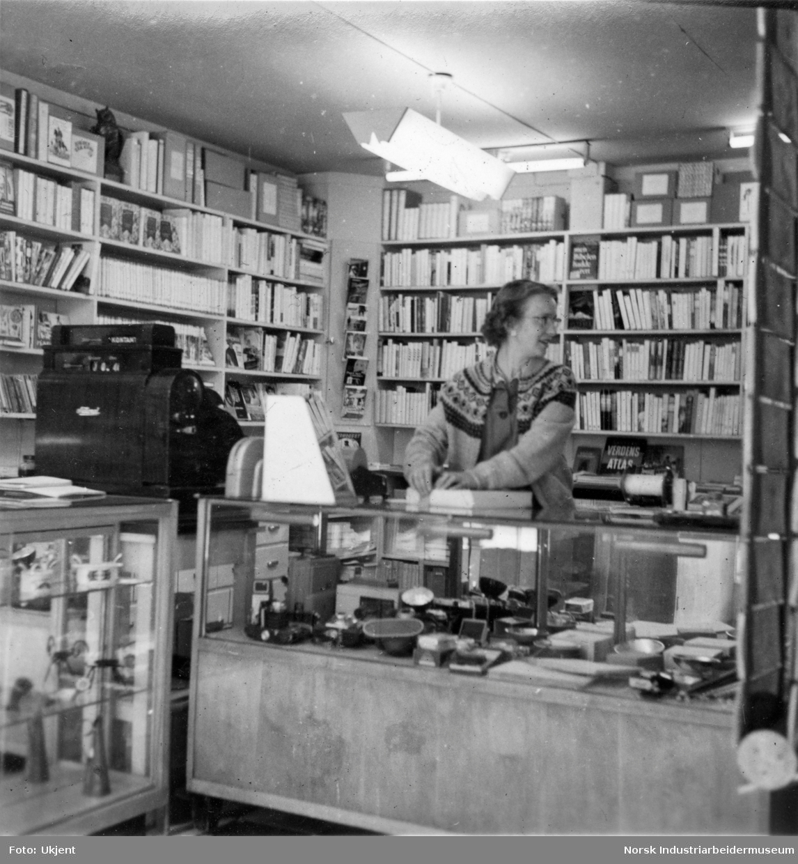 Butikklokale hos Køhn bokhandel og trykkeri, fotoavdelingen. Kvinne står bak disken med kasseapparat, bøker i hyllene på baksiden. Fotoutstyr i glassdisk.