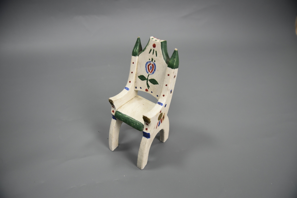 Stol til dokkemøblement i fem deler. Dette er en av tre identiske stoler. Den siste er litt mindre enn de andre. Møblene er hvitmalte med dekor i ulike farger. 
Møblementet er fra givers tidlige barndom, muligens 1928-1930. Møblene var til hennes første dokkestue, bygget av far av en appelsinkasse. 