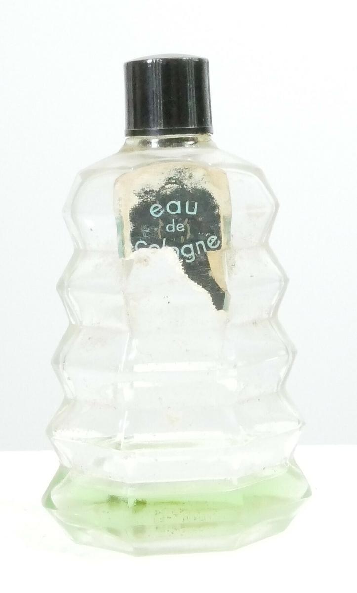 Pyramideformet glassflaske med svart rund plastkork og en påklistret papiretikett. Flasken inneholder grønn væske. 

Påskrift, etikett: 
eau // de // cologne 