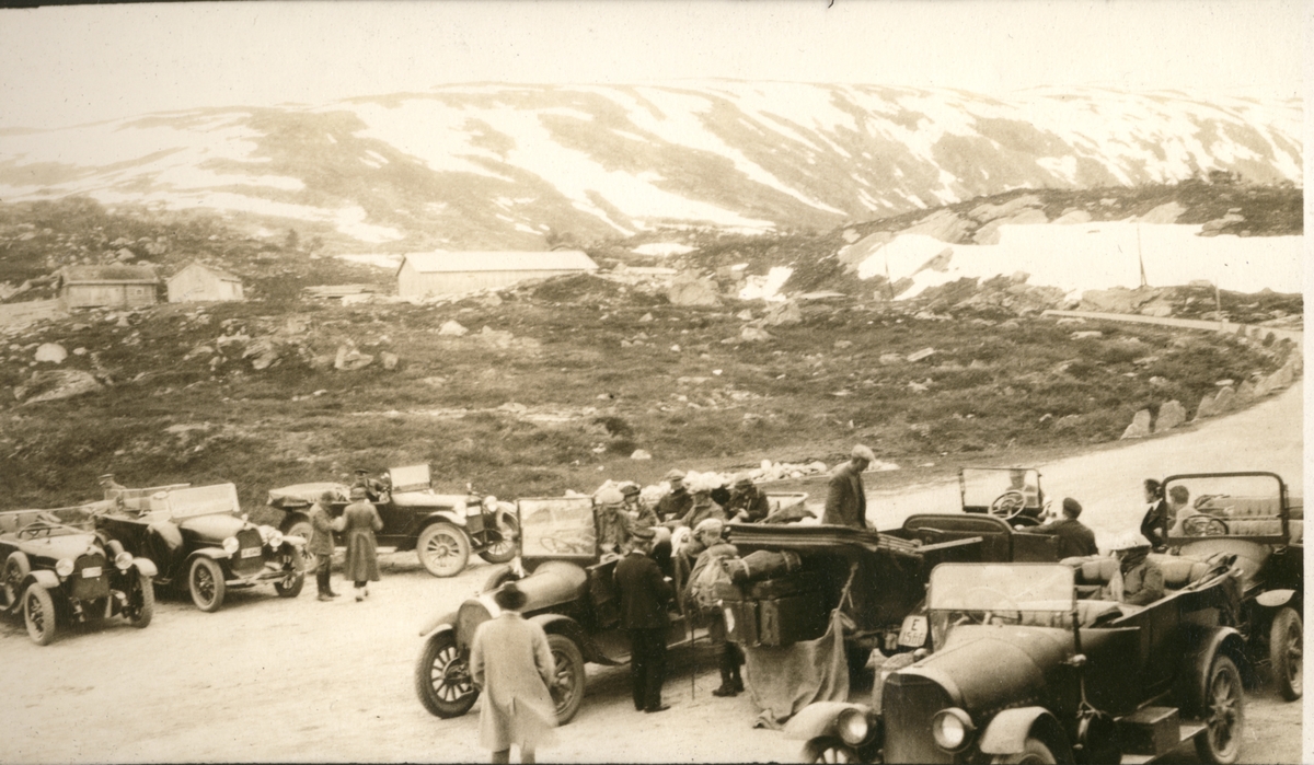 Syv biler(kabrioleter) med passasjerer har parkert på Grotli i Skjåk. I bakgrunnen et gårdstun. Fotografert juli 1923.