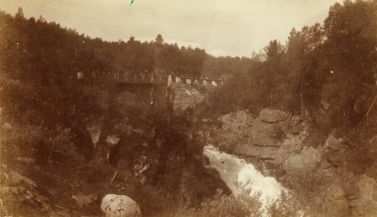 Søndenfjeldske Hjulrytterkompani sykler over bro ved fossefall, antagelig på vei mellom Koppang og Værnes for å delta i feltøvelser. Fofografert 1901.