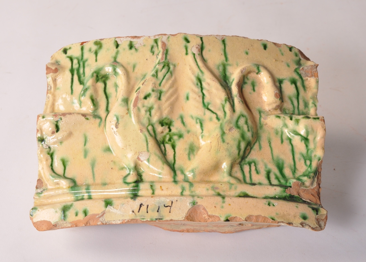 Kakelovn i deler fordelt i 16 trekasser av forskjellig størrelse, i tillegg 4 deler i pappeske. Deler av støpt kakel, glassert i gul og grønn med motiv av svaner.