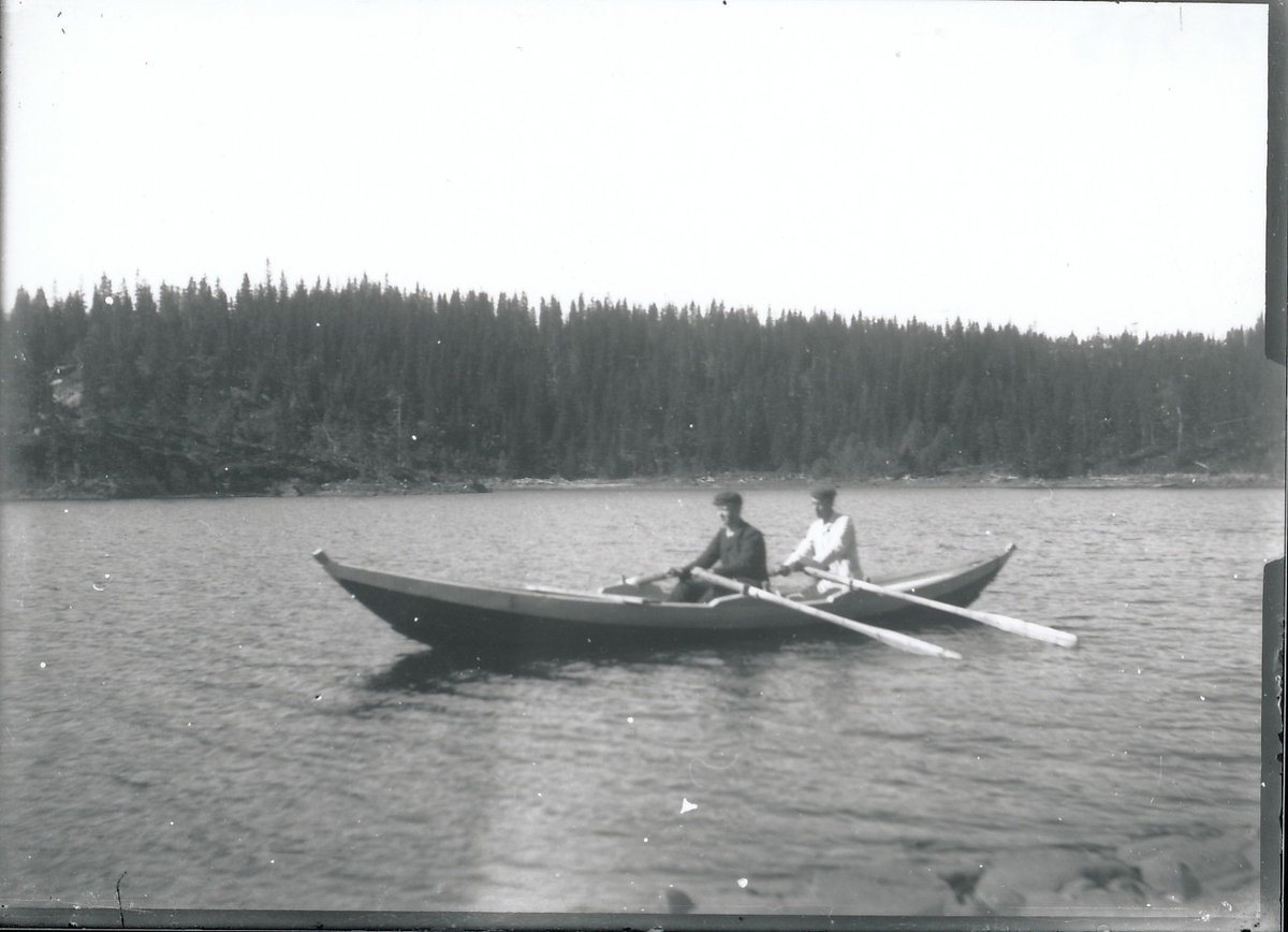 Harald Rydgren sitter forrest i båt og ror en trebåt på Mosjøen sammen med ukjent mann. Skog i bakgrunnen.
