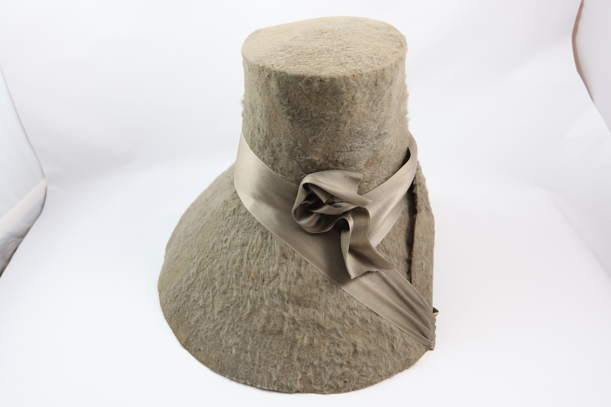 Stor grå kysehatt av filt/ull og kraftig brem forann sydd sammen. Tvers over sømmen og forlenget til hattebånd et grått silkebånd. Innvendig foret med grå silke.