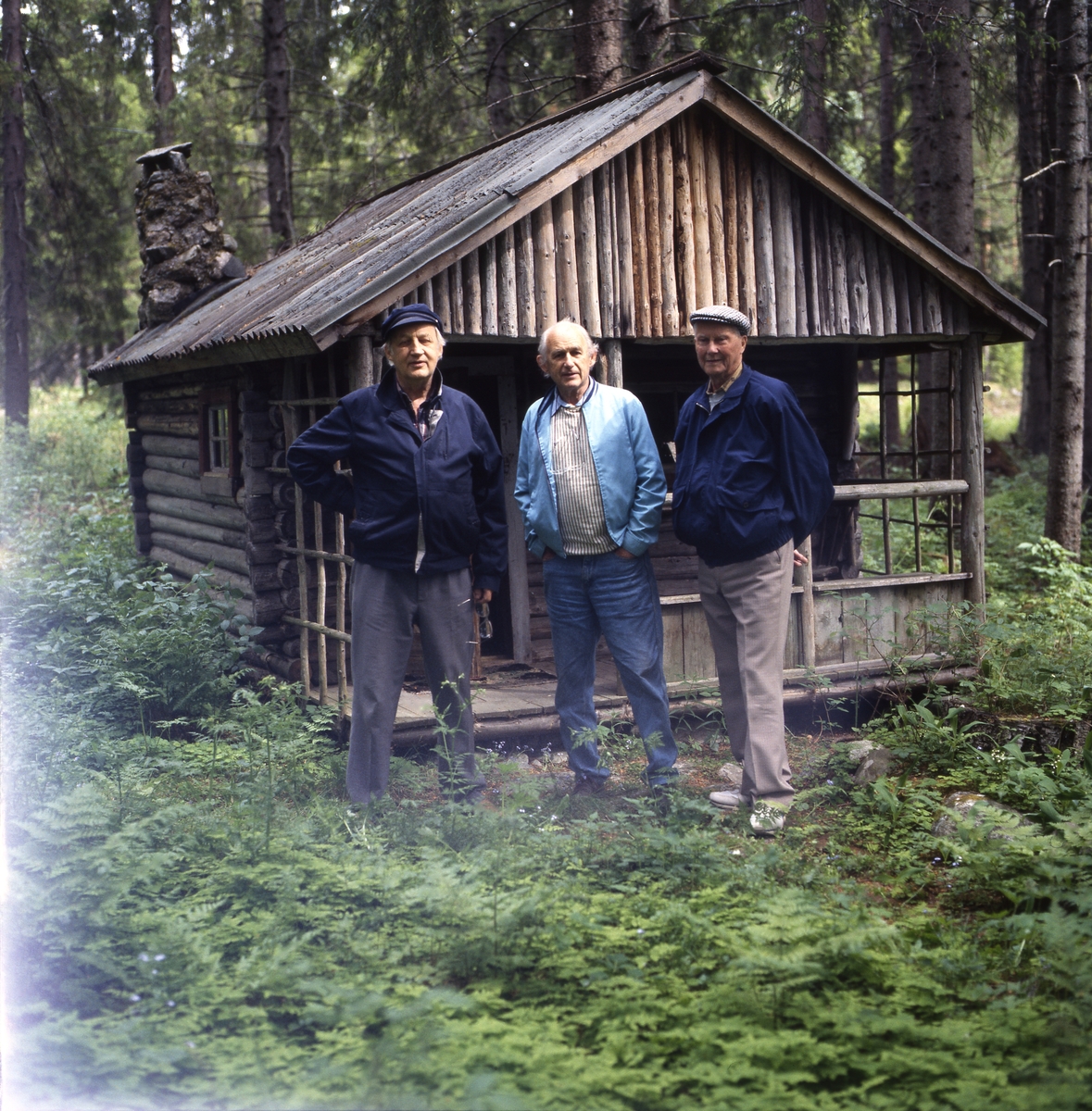 Några män, inklusive bröderna Ahnlund, står framför en gammal timmerstuga i en skogsglänta, 12 juni 1994.