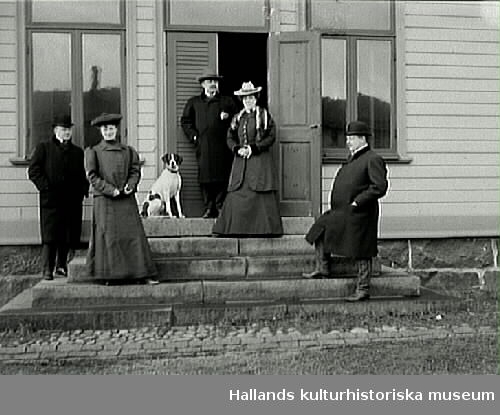 Tre män, två kvinnor och en hund på trappan till Himle härads tingshus. Ena dörrbladet står öppet och damen som står där bär en pälsstola.