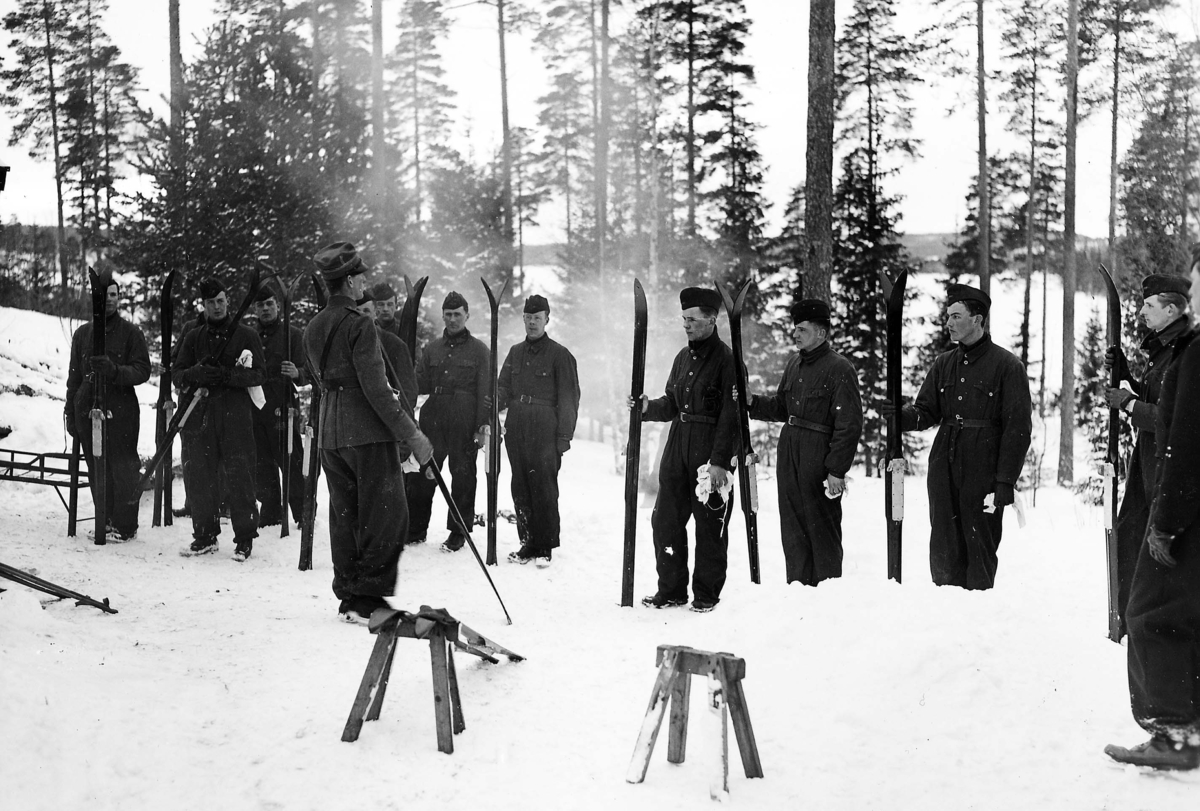 Flygsoldater står i en skog med skidor i hand, vintertid. Omkring tolv män tittar på en soldat. 1930-tal.