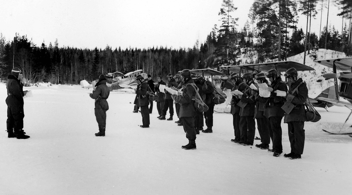 Ordergivning inför flygning vid vinterövning med F 5 Krigsflygskolan. Omkring 15 militära flygare i flygdräkt läser karta framför flygplan Sk 11, Tiger Moth. I bakgrunden syns flygplan Sk 6. Cirka 1932-1939.