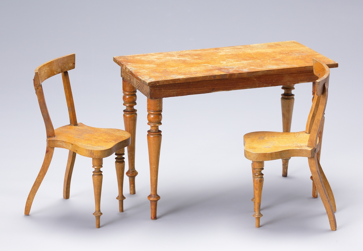 Två dockstolar i trä med formsågad sits, svängt ryggstöd och svarvade ben framtill. Stolarna ingår i ett möblemang med ett tillhörande bord.