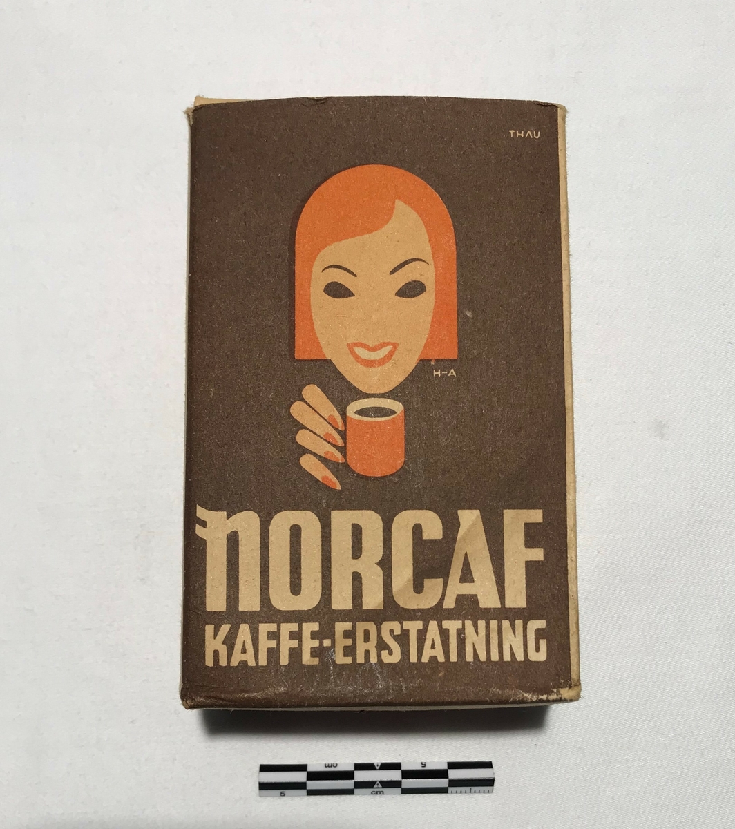 Eske av papp med kaffeerstatning. Tekst: NORCAF KAFFE ERSTATNING. På sidene står bruksanvisning på tilbereding for koking og trakting. Kaffeerstatningen ligger i esken i en papirpose.