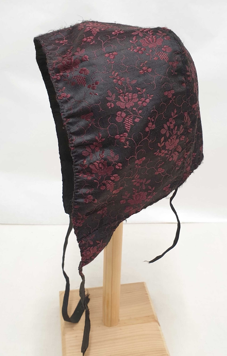 Sydd av svart, blomstret silkedamask med rødfiolette blomster. Fóret med svart linstoff, ripsaktige ca. 1,3 cm brede knytebånd Dobbel rad stikninger i kanten, glatt bakstykke.