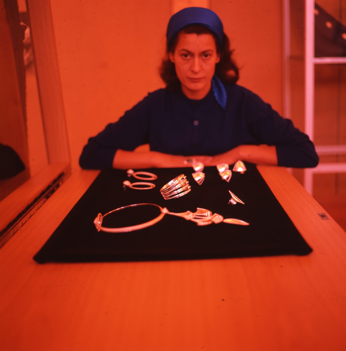Silversmeden Rosa Taikon sitter vid ett bord där hennes silversmycken ligger upplagda på en skärm. Bilden är tagen i samband med hennes debututställning på Bollmora bibliotek år 1966.