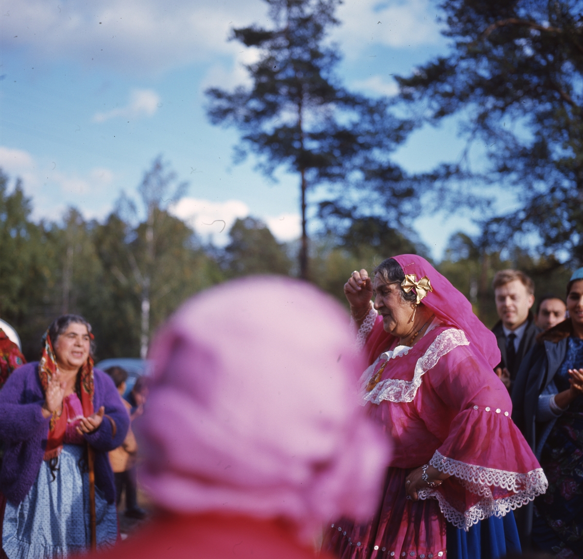 Bilden är troligen tagen i Skarpnäckslägret, Stockholm där man samlats för att se den rysk-romska artisten Raya Udovikova, verksam vid Romén-teatern i Moskva.  En snarlik bild finns publicerad i tidskriften Zigenaren från 1966, tagen i samband med att Udovikova gästade Sverige. Hon gjorde då ett besök i Skarpnäckslägret och på bilderna i tidskriften syns Udovikova dansa tillsammans med boende i lägret. I bildtexten uppges att Udovikova  tillsammans med en av de boende i lägret sjöng gamla romska sånger, ackompanjerade på dragspel.