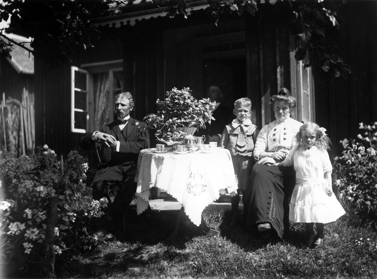 Två män och en kvinna med två barn: en pojke och en flicka, vid dukat bord i trädgården.