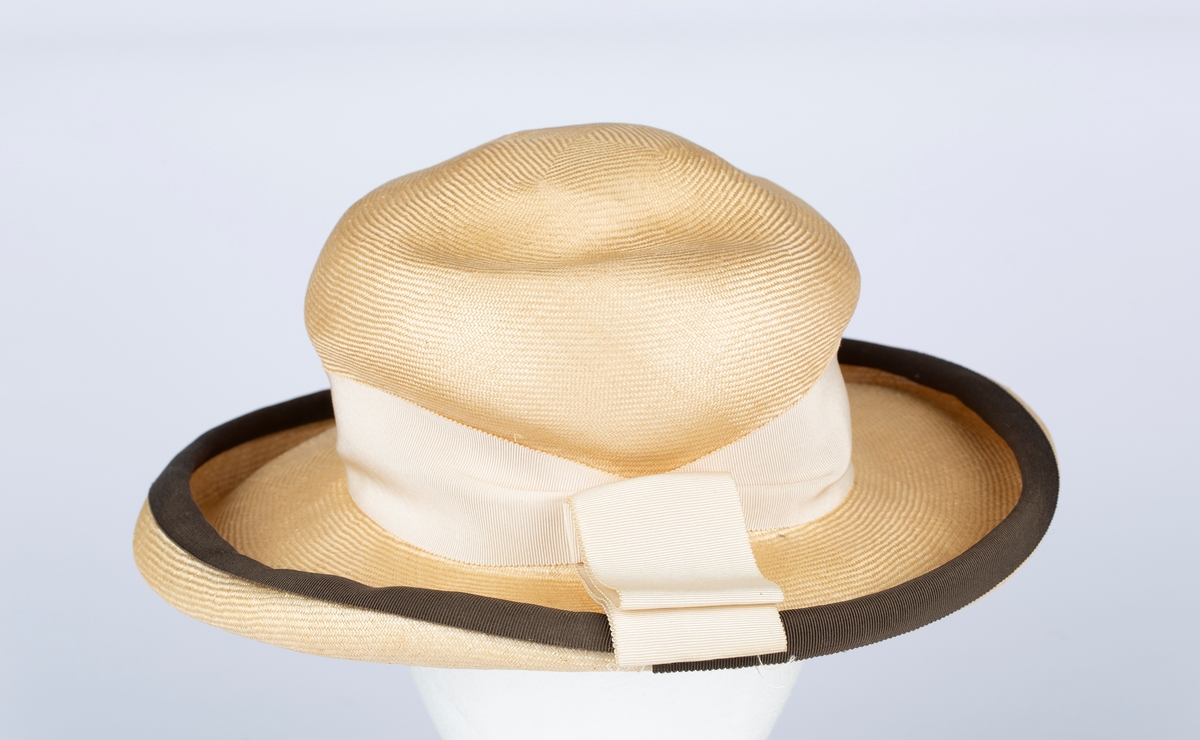 Rund hattepull, vid brem kantet med brunt ripsbåndhattebånd. Rundt hodepullen bredt ripsvevet , lyst hattebånd med sløyfe bak.