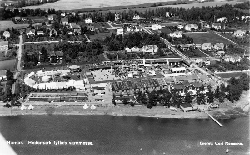 Flyfoto som viser tivolitelt til venstre og en stor plass med utstillingsboder samt Ridehuset på Hamar og omliggende villabebyggelse. Mjøsa i forkant. (Foto/Photo)