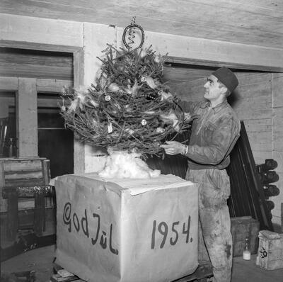 Svart-hvitt bilde av lite juletre som står på en firkantet boks påskrevet "God jul 1954". Til høyre for treet står en mann i kjeledress. Juletreet er pyntet med metallspiraler mm og befinner seg i Hamar jernstøperis lokaler.. Foto/Photo