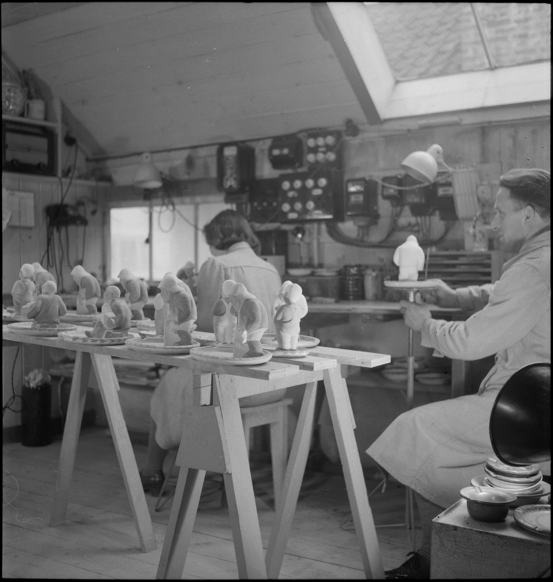 Keramiker Thorvald Olsen og en kvinne maler keramiske produkter i Olsens verksted. På bordet er det flere keramikkfigurer.