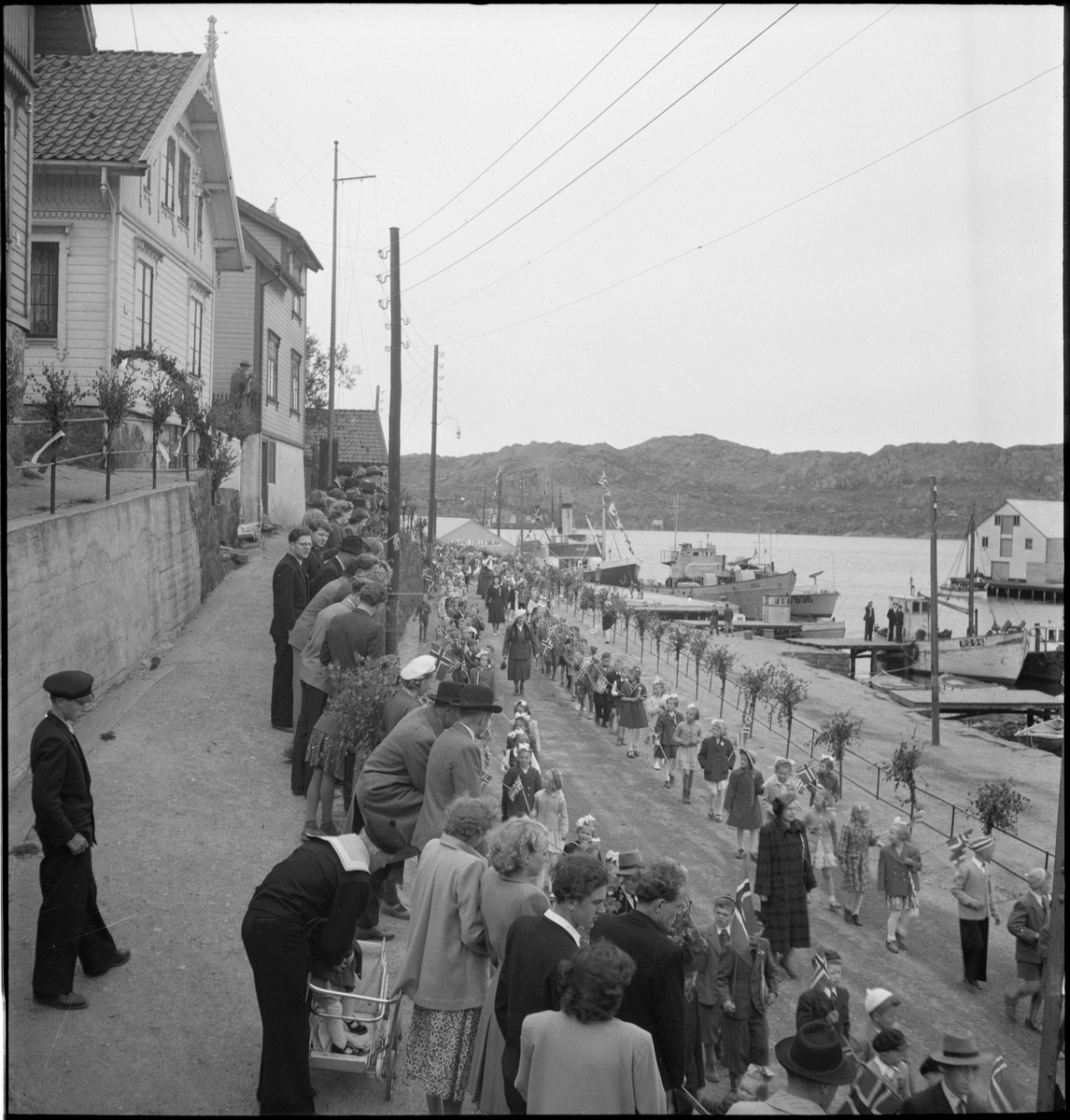 Folketog på 17. mai. Flere korps og barn går langs Vågen og igjennom sentrum av Egersund. Mange har kommet for å se toget.
