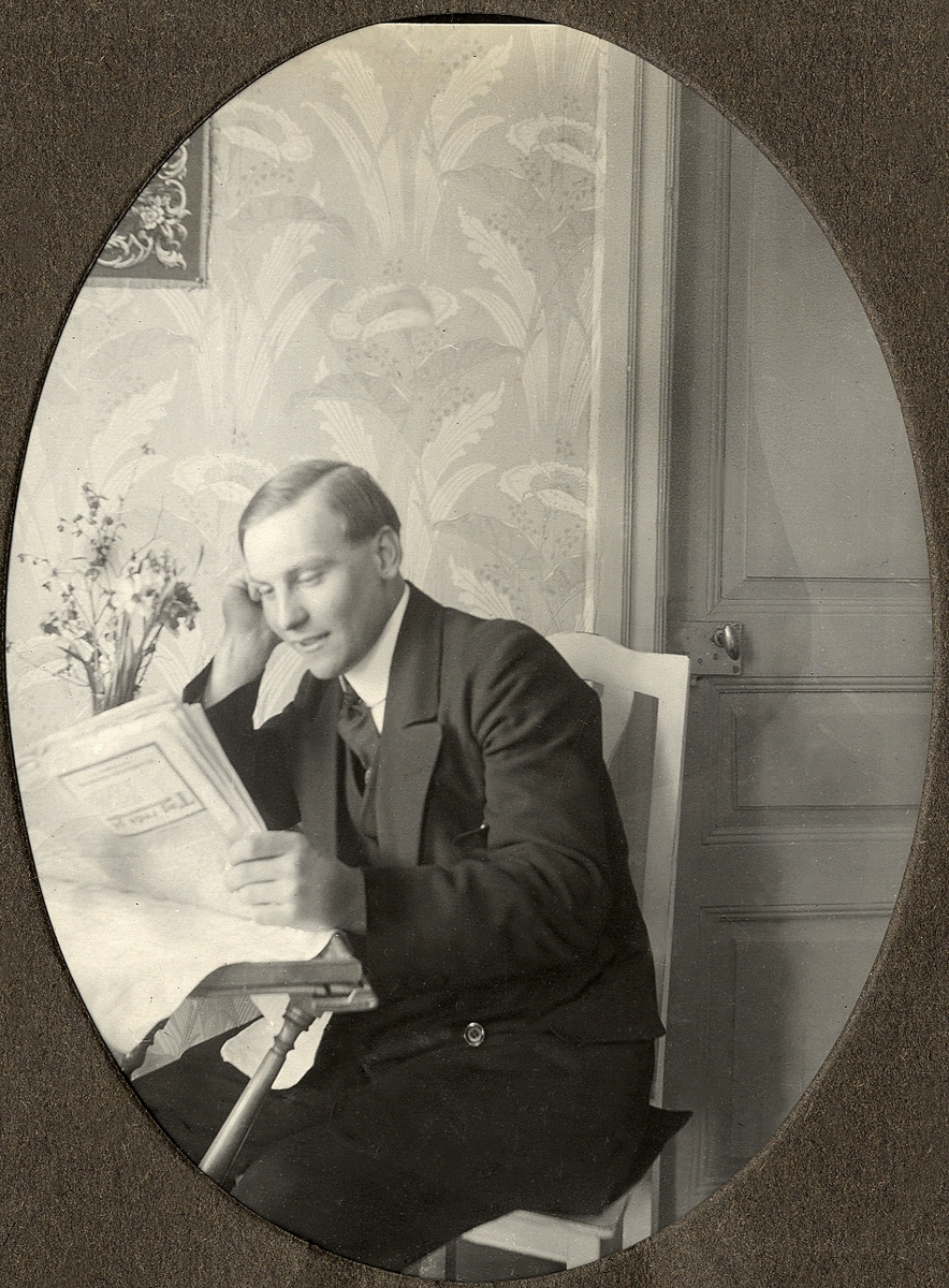 En ung man i kostym sitter och läser tidningen vid ett bord.