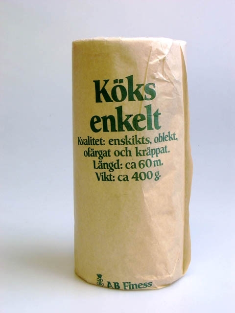 En pappersförpackning med en rulle hushållspapper. Grön text: "Köksenkelt, Kvalitet: enskikts, oblekt, ofärgat och kräppat. Längd:ca 60 m. Vikt: ca 400 g. AB Finess". Förpackningen har införskaffats till en utställning på Landskrona museum 1990, som hette "Papper, papper, papper".