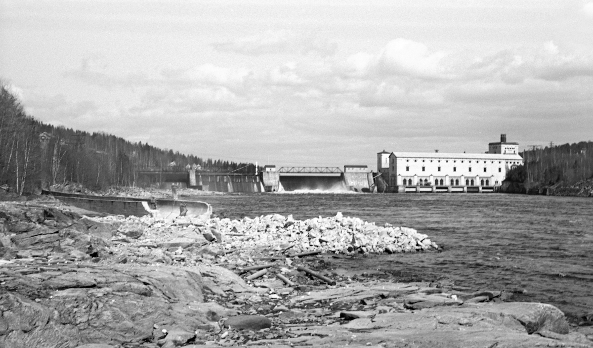 Fra rivinga av tømmerrenna ved Rånåsfoss i Glomma, nærmere bestemt i Sørum i Akershus, i 1962. Dette fotografiet viser dammen og kraftstasjonen i motstrøms retning. Anlegget ble bygd i perioden 1918-1922 på oppdrag fra Akershus fylke. Bygningene ble tegnet av den kjente arkitekten Thorvald Astrup (1876-1940). Dammen er 186 meter lang og 18 meter høy. Den ble bygd med et isløp nærmest kraftstasjonen og to 50 meter brede sektorløp, samt et 45 meter bredt valseløp som skulle brukes til tapping under flom og i vintersesongen, da sektorlukene sto i faste posisjoner. Sommerstid var sektorlukene driftssikre og enkle å manøvrere. I utgangspunktet ble størstedelen av tømmeret ledet gjennom disse, som kunne ta unna opptil 700 000 stokker i døgnet med relativt beskjedent vannforbruk. I tillegg ble det støpt ei 800 meter lang åpen tømmerrenne ved den vestre elvebredden. Den skulle brukes når vannføringa i elva var under 450 meter i sekundet. Under slike forhold kunne renna ta unna opptil 180 000 stokker i døgnet med et vannforbruk på 15-30 kubikkmeter i sekundet. Renna var svært sjelden i bruk, og når den var i bruk krevde tømmerekspedisjonen et betydelig mannskap til påstikking. Dette kan ha vært bakgrunnen for at renna ble fjernet. I begynnelsen av 1980-åra ble det bygd et nytt kraftverk - Rånåsfoss II - der hvor tømmerrenna hadde gått.