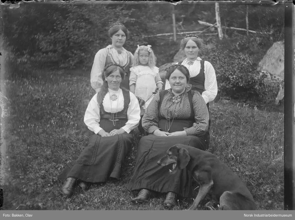 Fire kvinner sittende utendørs i bunad sammen med jente i hvit kjole. Hund sittende på siden.