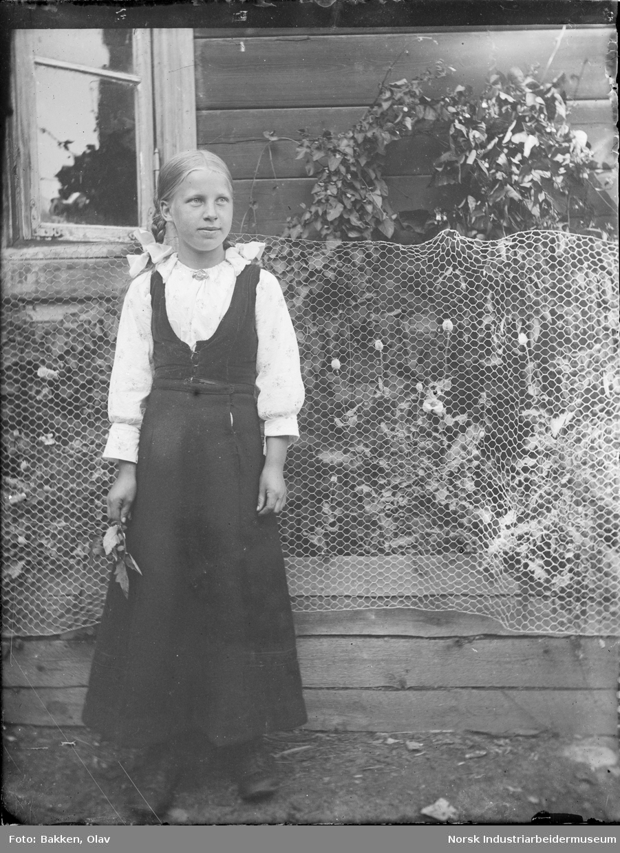 Portrett. Jente stående foran bygg med blomst i hånden. Fletter og sløyfer i håret, kjole. Netting langs veggen på huset i bakgrunnen.