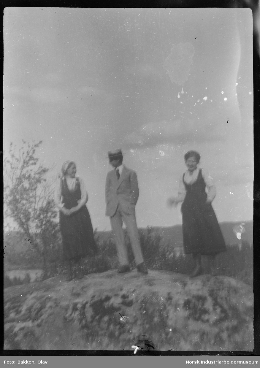 Mann og to damer stående på fjellknaus. Mannen i stråhatt og dress, kvinnene i drakt.