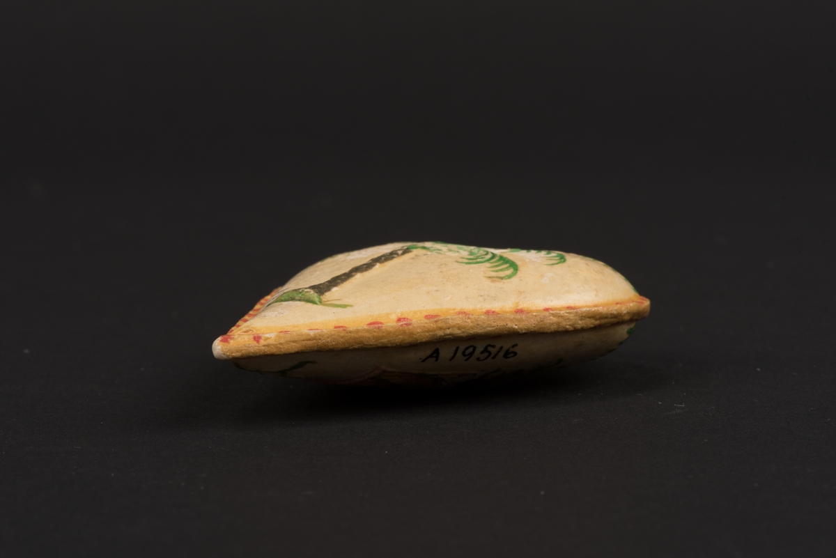 Kotiljongsmärke, troligen av papier-maché, i form av ett stoppat hjärta.
Hjärtat är dekorerat på bägge sidorna och har en kant målad i gult och rött. På den ena sidan avbildas en randig blomsterkorg som innehåller gröna blad och röda hjärtan under en girlang. På andra sidan är en palm under girlang. På denna sida finns även en inskrift i blyerts: Söderköping 1850.