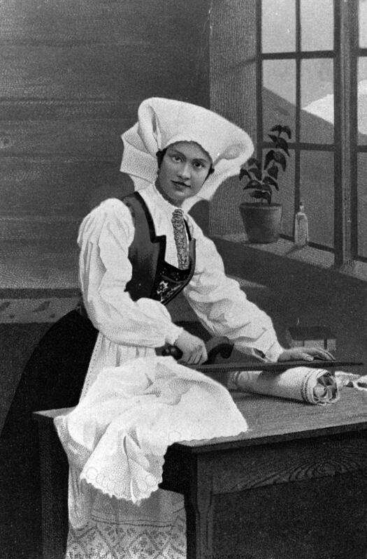 Svart-hvitt foto av kvinne i bunad med stort hodeplagg, som bruker et mangletre til å glatte tekstiler.