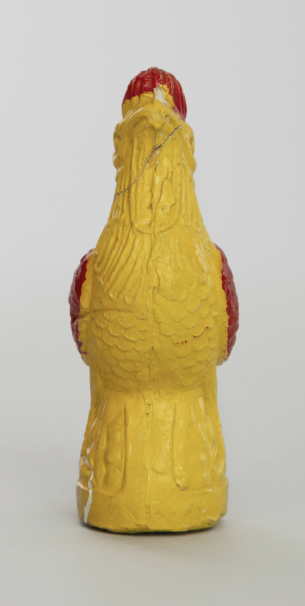 Spaltefløyte av keramikk. Formet som en hane. Støpt i to halvdeler. Malt gul med røde vinger og kam.
Spalte og labium i stjerten. Ett fingerhull.