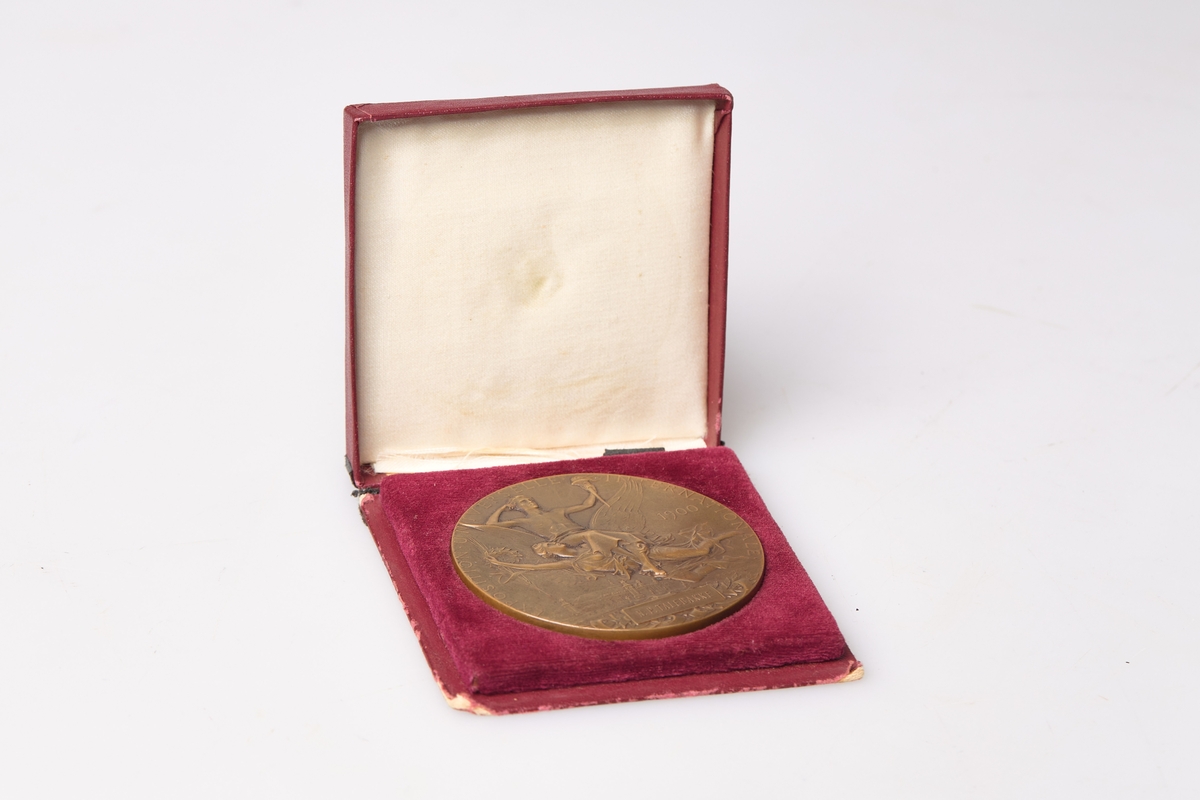 Medalje og rødt etui. Den bronsestøpte medaljen med gravert innskripsjon er datert til 1900. Medaljen er produsert i Frankrike og er en pris gitt på utstilling.