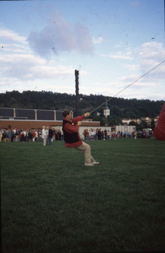 En man står bakåtlutad och drar i en lina till en luftballong. I bakgrunden står en skara männiksor och tittar mot en stigande luftballong som inte synd i bild.