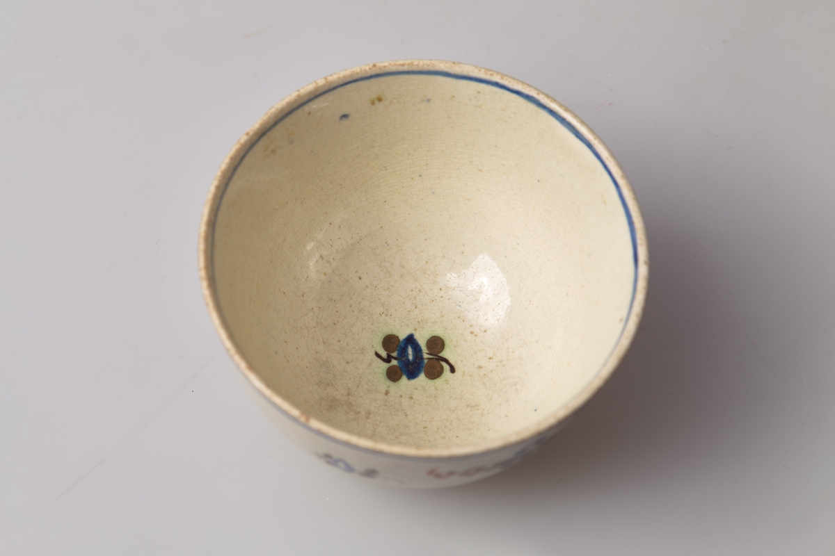 Kopp uten hank i glasert steingods. Håndmalt blomstermotiv i blått, brunt og grønt, samt en blå ring på både inner- og yttersiden av øvre kant. 
