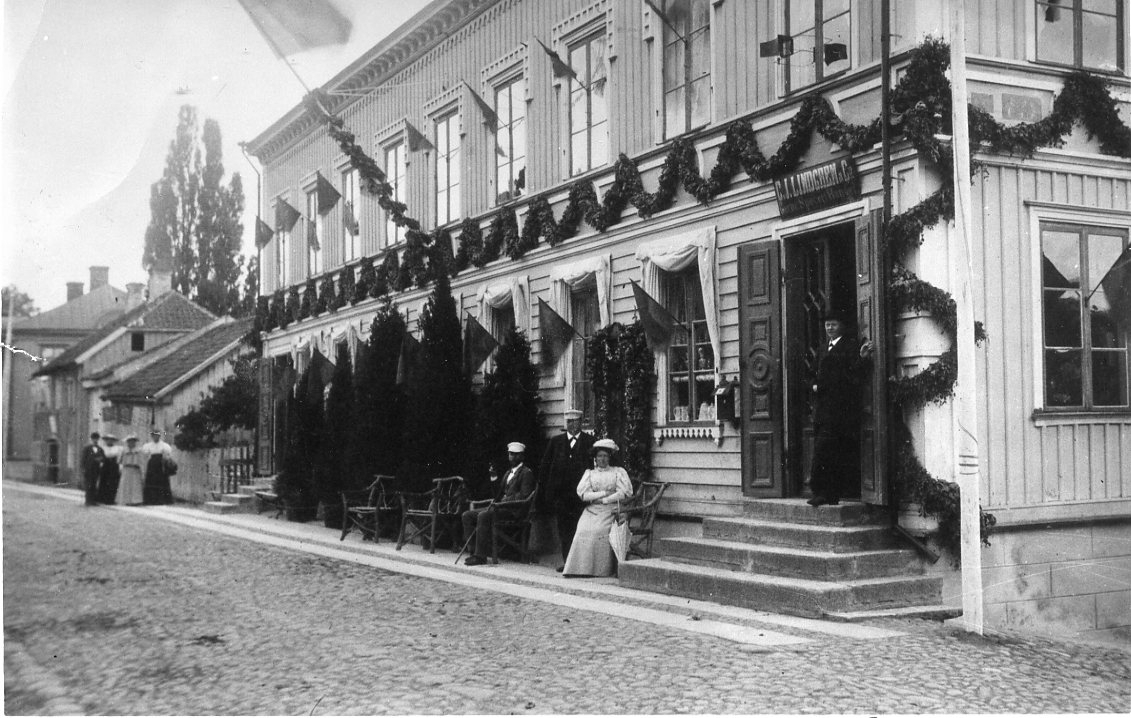 Ett dekorerat med blomstergirlanger och hissad fasadflagg. Utanför sitter och står tre personer, vid entrén till C. J. Lindgren & Co. står en man. Längre bort i bild kommer tre kvinnor och en man promenerade i riktning mot fotografen. De väntar på kung Oscar II.