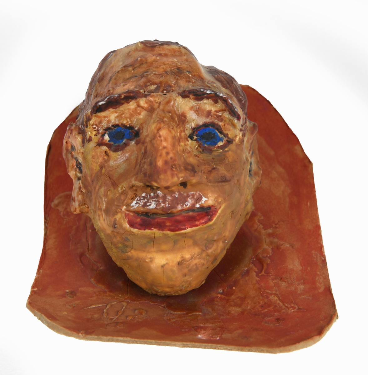 Et mannshode av papp/papir laget av en elev (i formingsfag). Hodet er i pappmasje og er montert på en brunmalt papplate. Hodet er malt i brunt med mørke brune øyenbryn, blå øyne og rød munn.