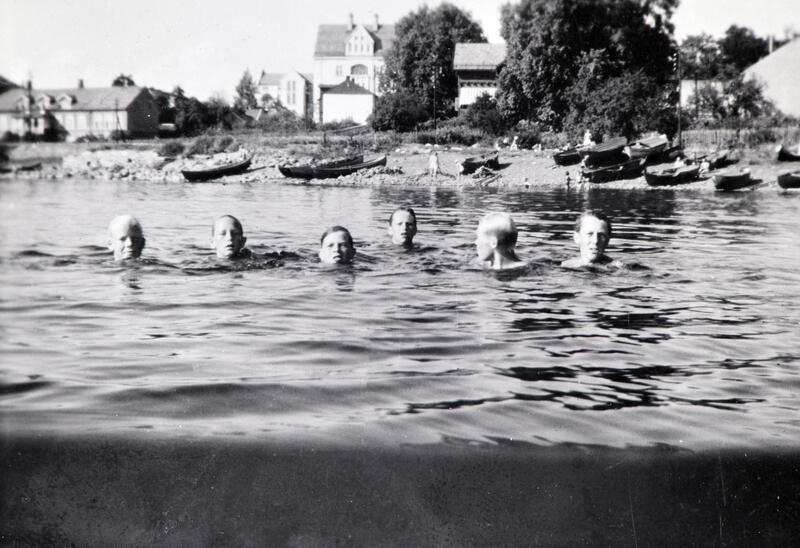 Svart-hvitt foto av seks mannshoder som stikker opp av vannet under en svømmetur. Bymiljø i bakgrunnen. Koigen, Hamar.