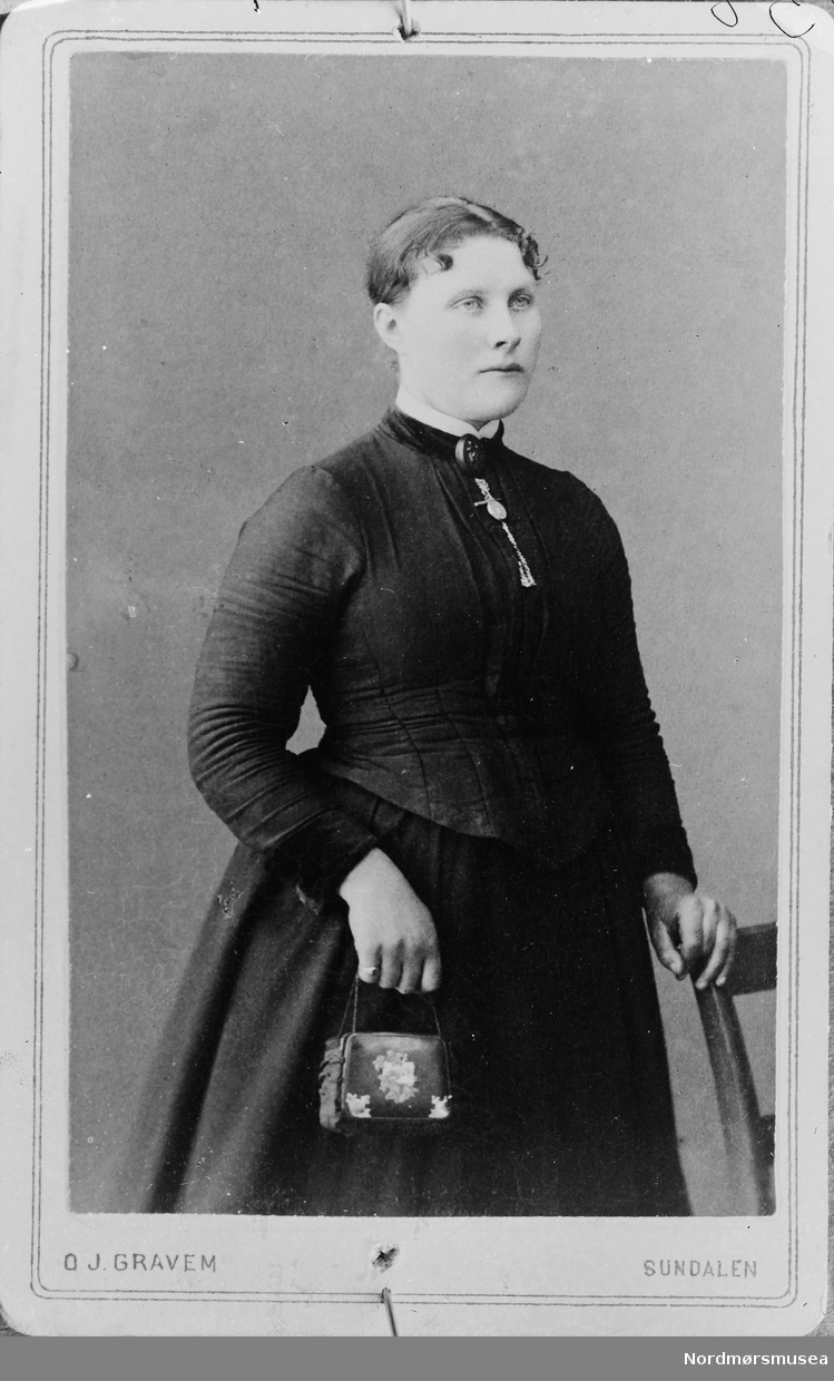 Bilde av en av fotografens framkalte bilder. Er negativet blitt borte? Stående kvinne i mørk kjole med veske i ene hånden og andre hånden på en stol.